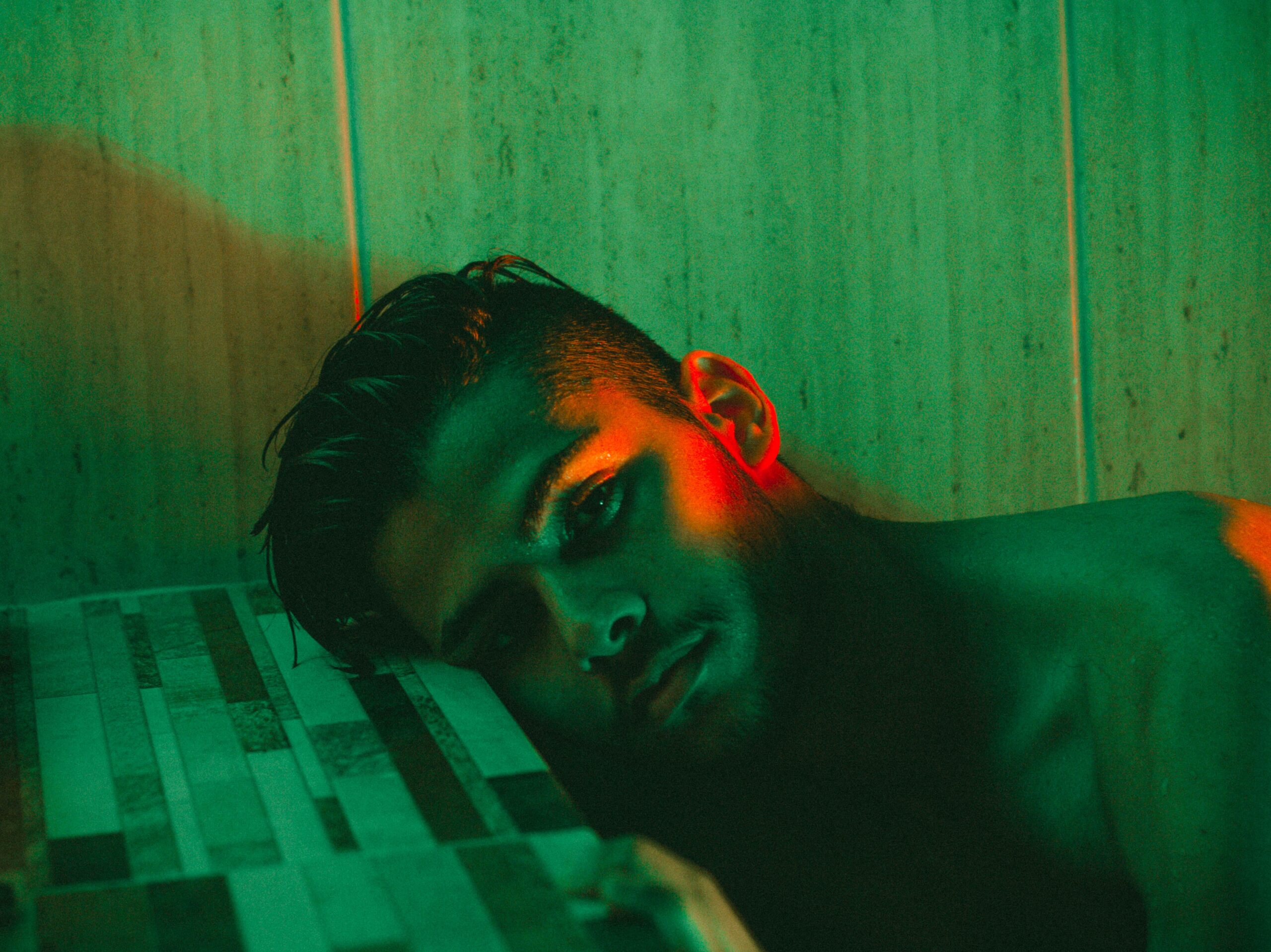 A shirtless person with wet hair lies with their head on a tiled surface, illuminated by dramatic green and red lighting against a tiled wall—capturing the vulnerability of finding human connection in an urban solitude.