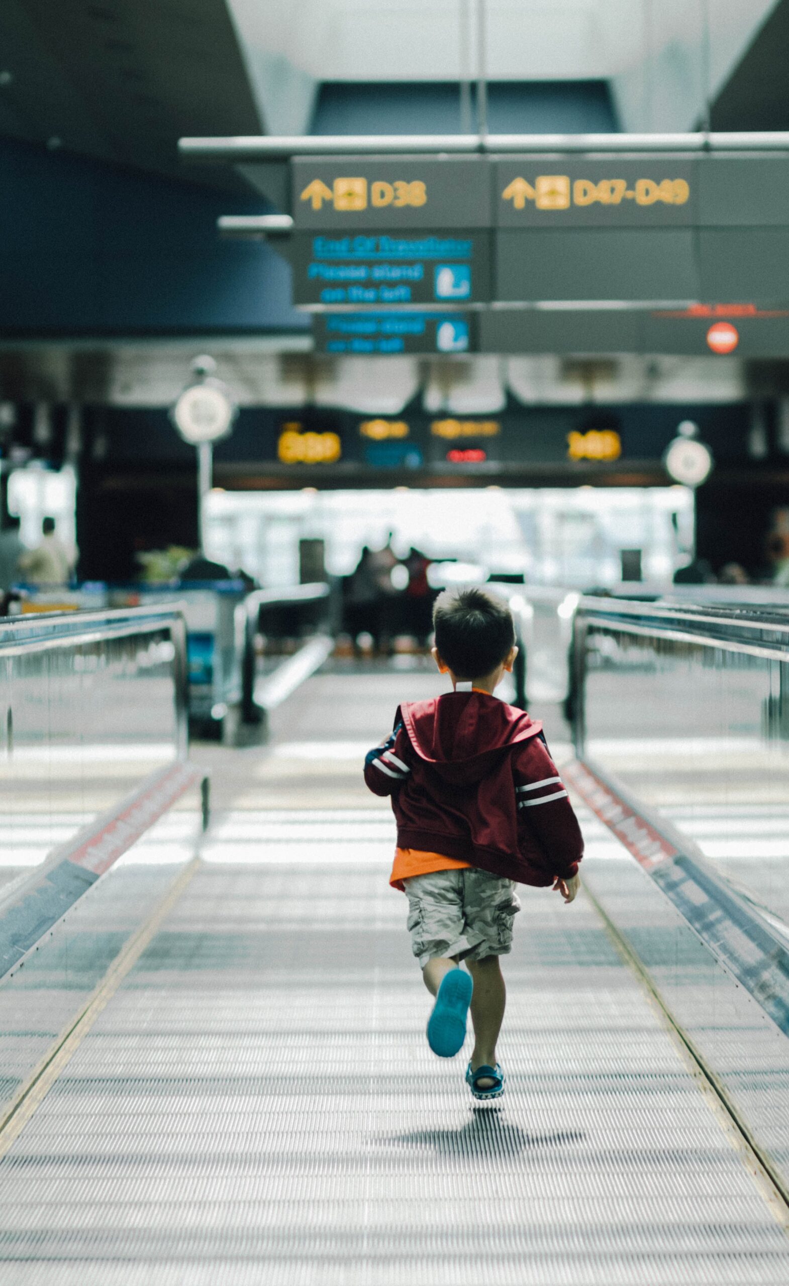 A young child in a red jacket walks alone on a moving walkway inside an airport, embodying the journey of embracing failure and growth, with signs for gates and blurry travelers visible in the background.