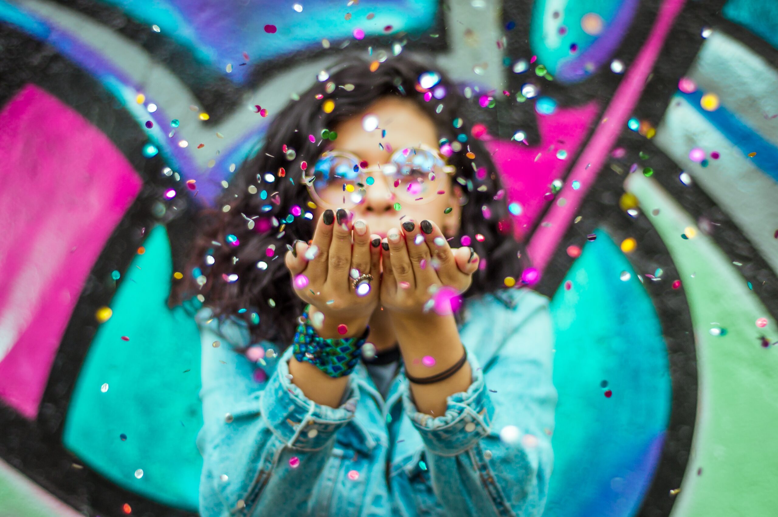 A person wearing glasses and a denim jacket blows colorful confetti toward the camera, with a vibrant graffiti wall in the background—perfect inspiration to make your own free Father's Day card.