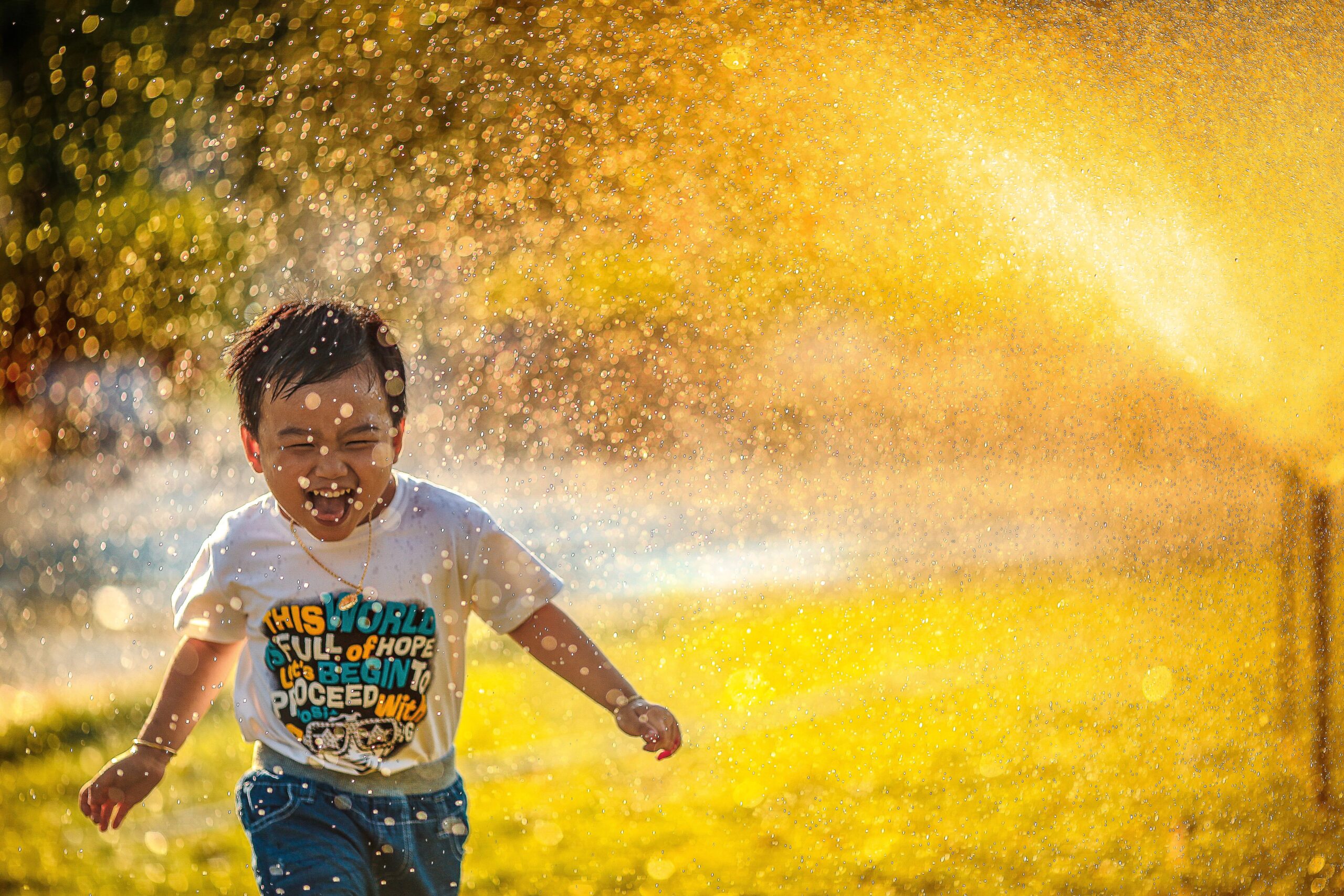 A young child laughs joyfully while running through a spray of water outdoors, sunlight shining through the droplets like meditation hacks for the soul. The child wears a white t-shirt and blue shorts, every moment free from wasted efforts.