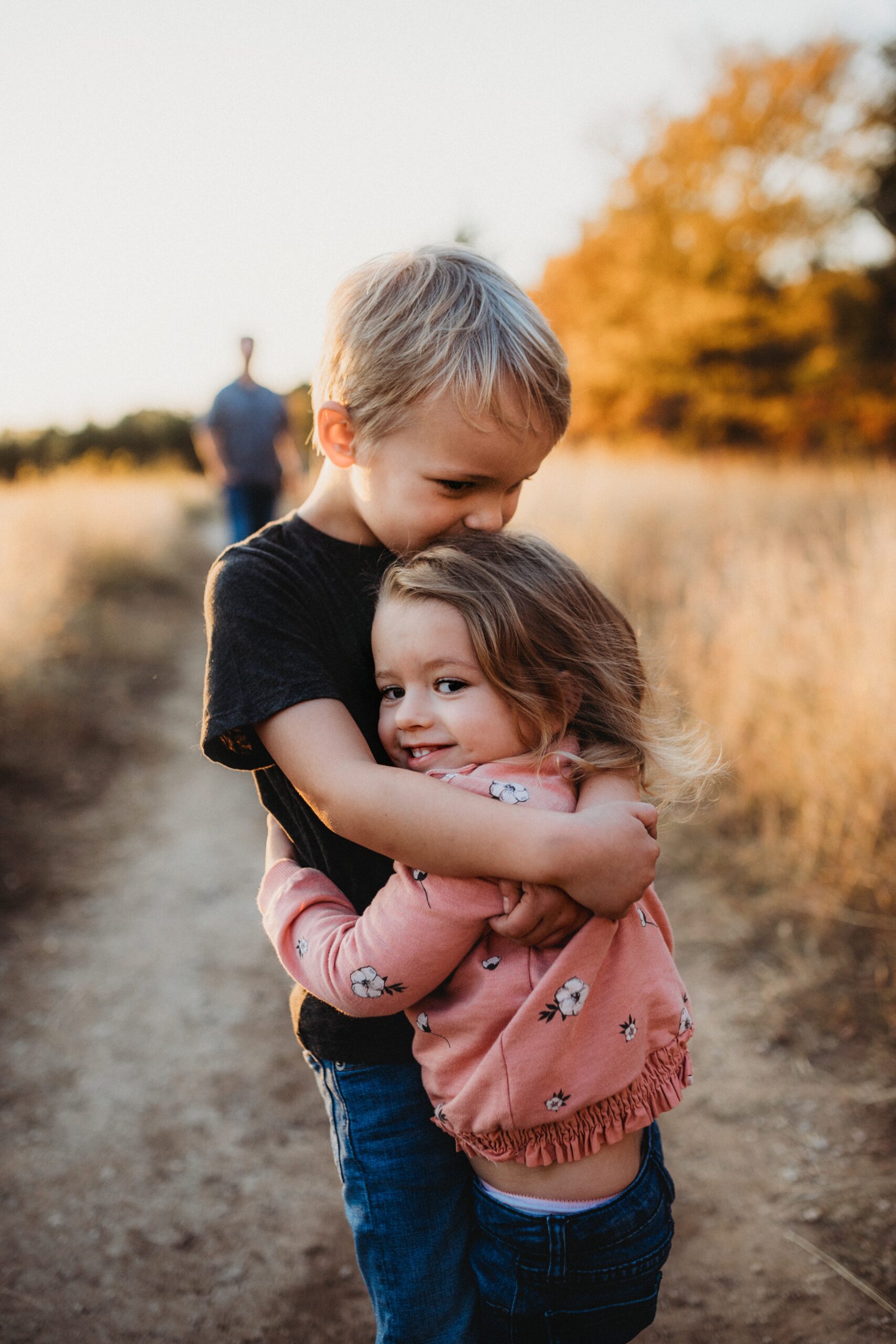 A young boy and a little girl hug each other on a dirt path through tall grass, embodying the power of apology. The girl smiles at the camera while the boy kisses her head, as an adult walks in the blurred background under warm evening light.