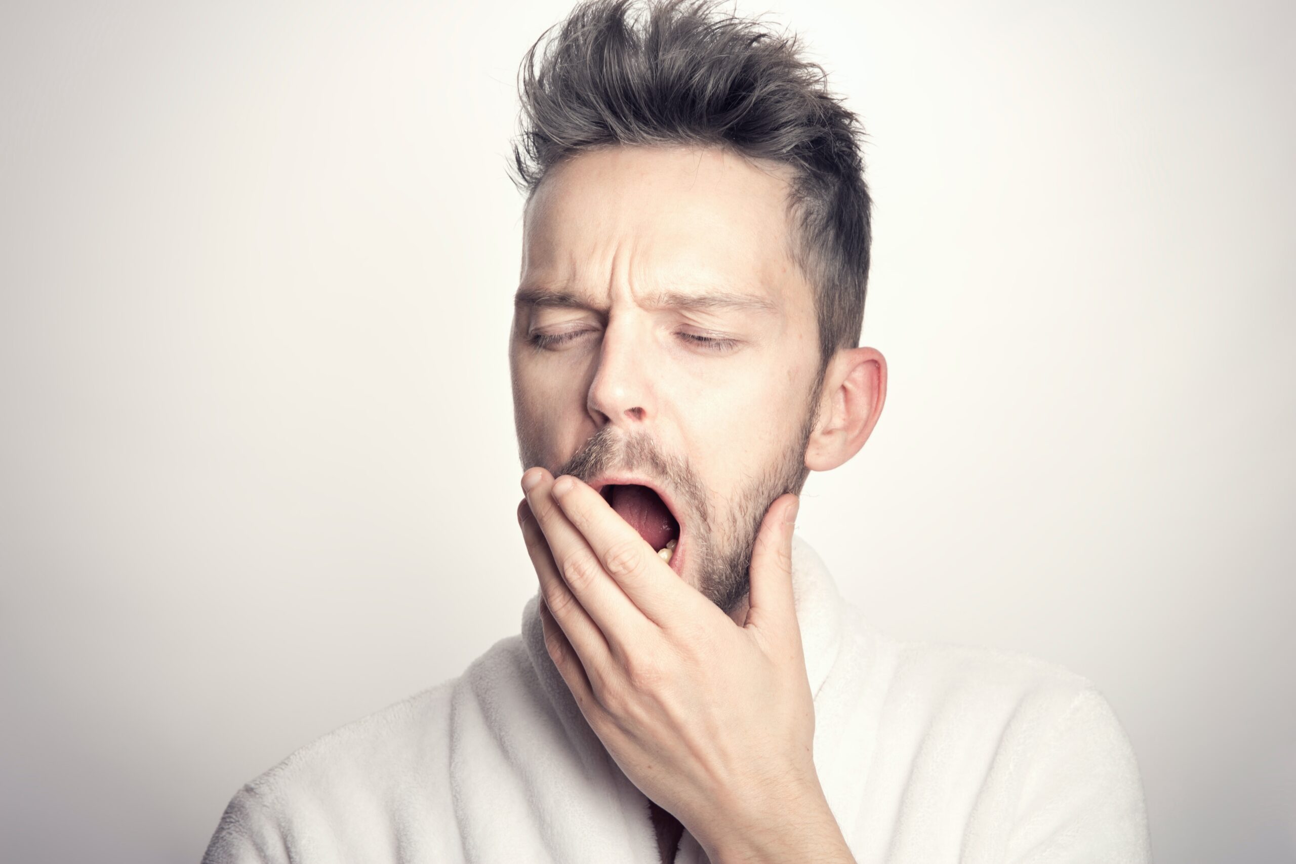 A man wearing a white robe is yawning with his eyes closed and one hand covering his mouth, embodying the calm of restful meditation and the luxury of time wealth against a plain light background.