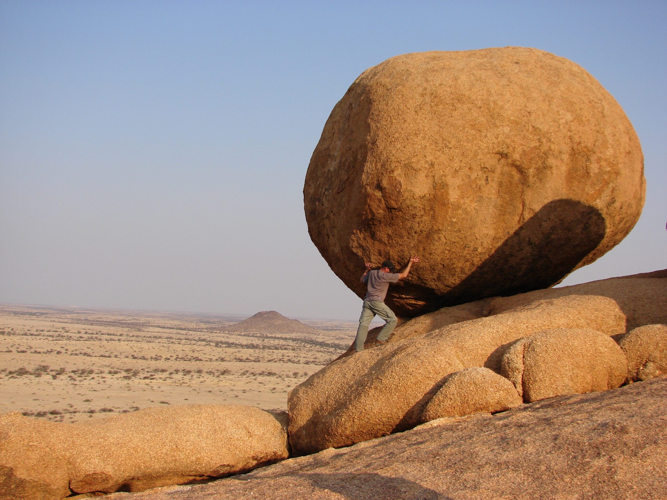 A person appears to push a massive round boulder atop rocky terrain in a wide, arid landscape under a clear sky, symbolizing the challenge of managing change amidst difficult conditions.