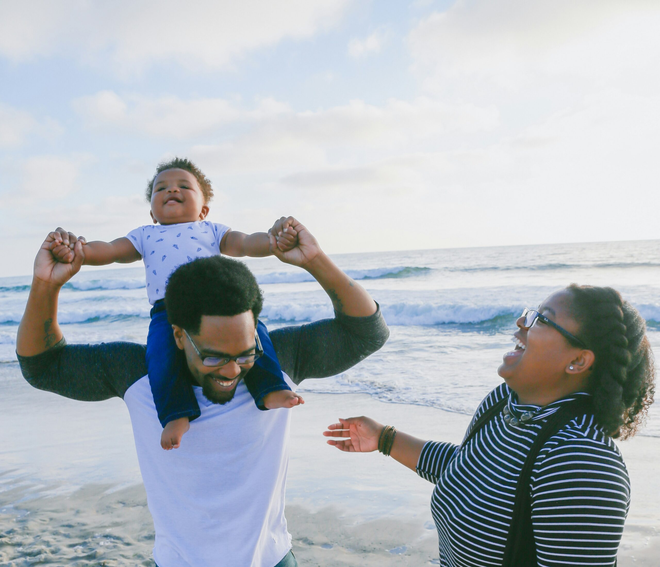 A smiling man holds a happy toddler on his shoulders while a woman laughs beside them; they are standing on a beach with waves and a bright sky in the background.