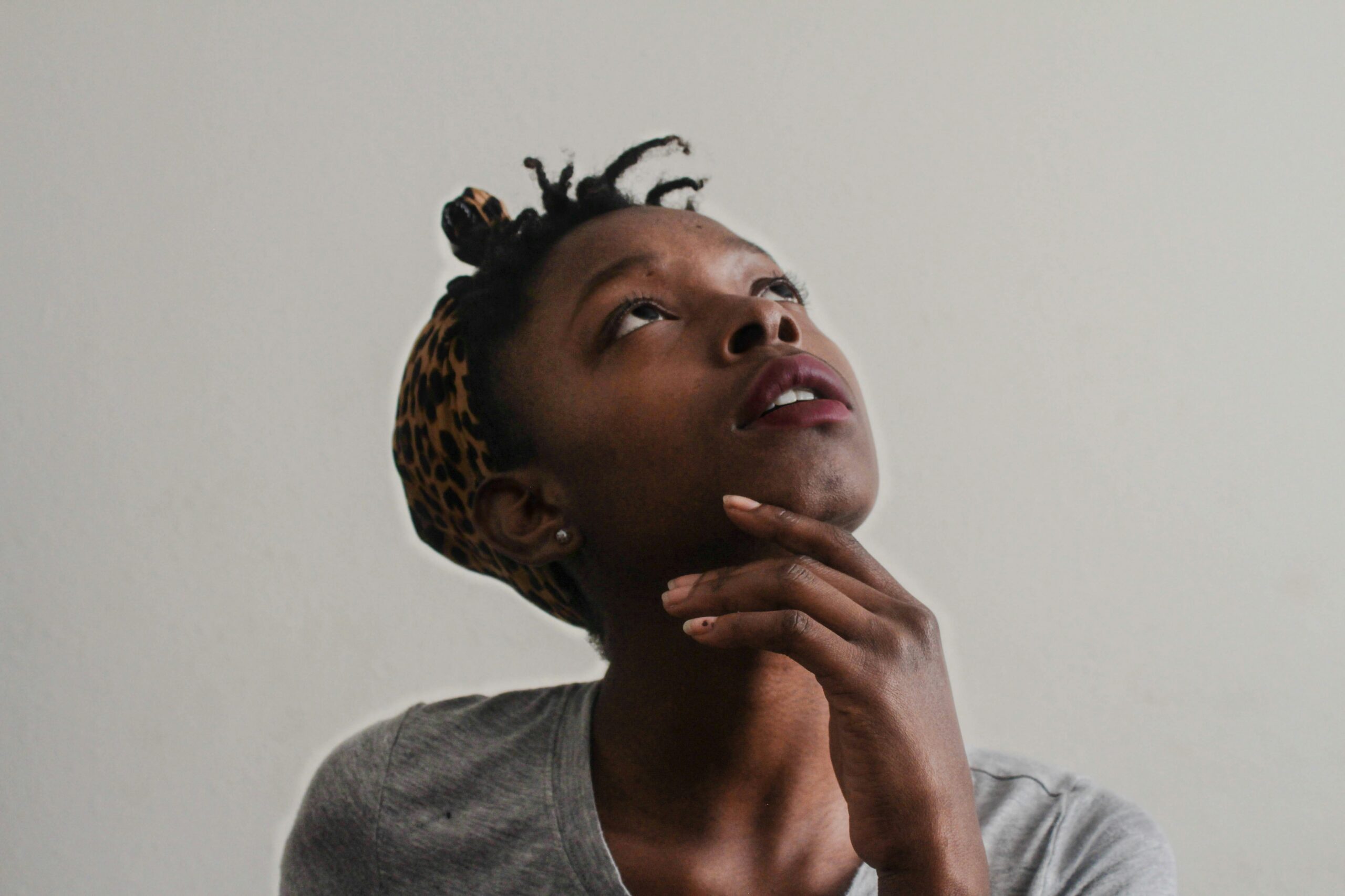 A person wearing a gray shirt and a leopard print headband looks thoughtfully upward, resting their chin on their hand against a plain light background, as if reflecting on managing family responsibilities and mental load.