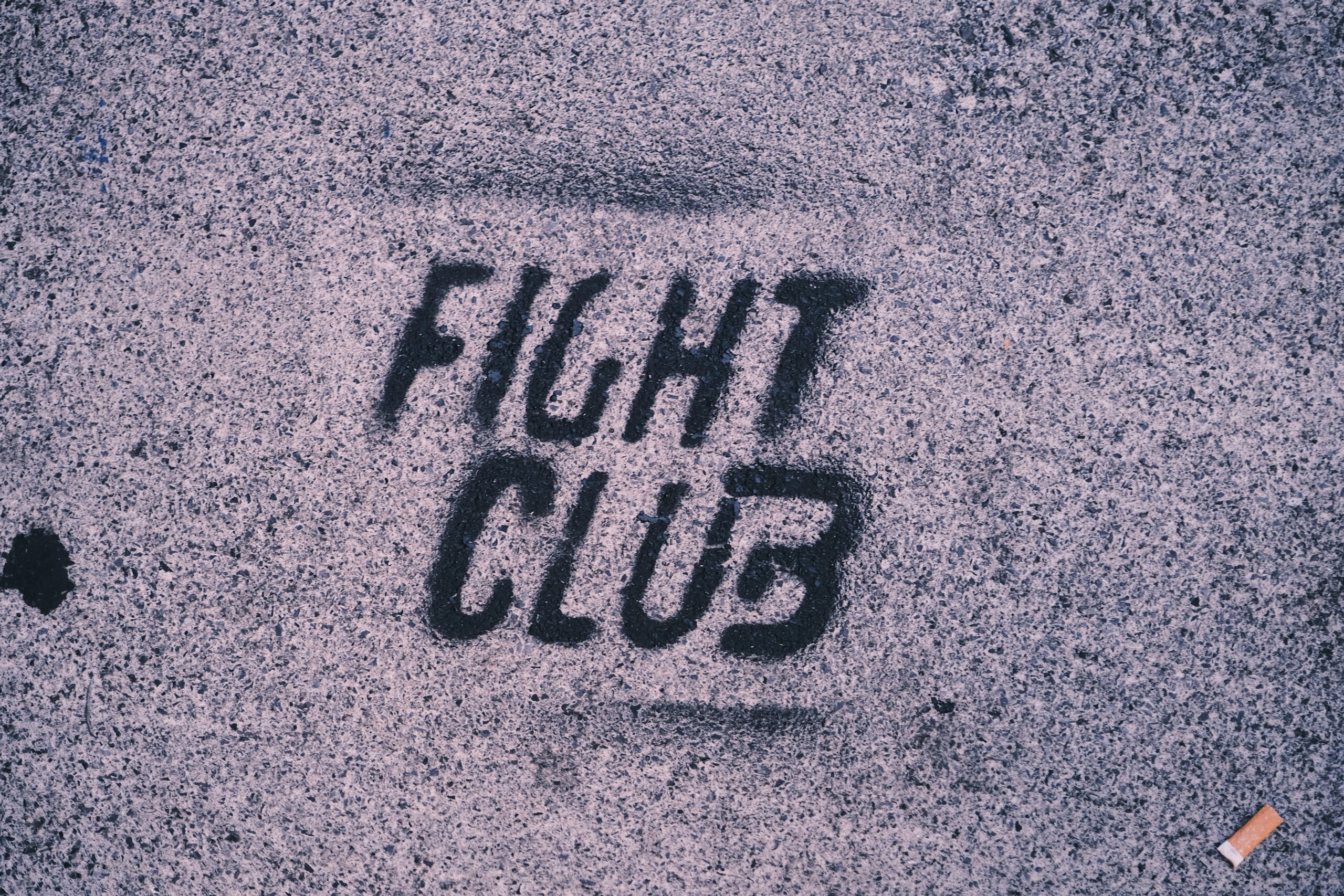 Black stencil graffiti reading FIGHT CLUB is painted on a rough gray concrete surface, evoking themes of navigating masculinity. A small cigarette butt lies on the ground in the bottom right corner.