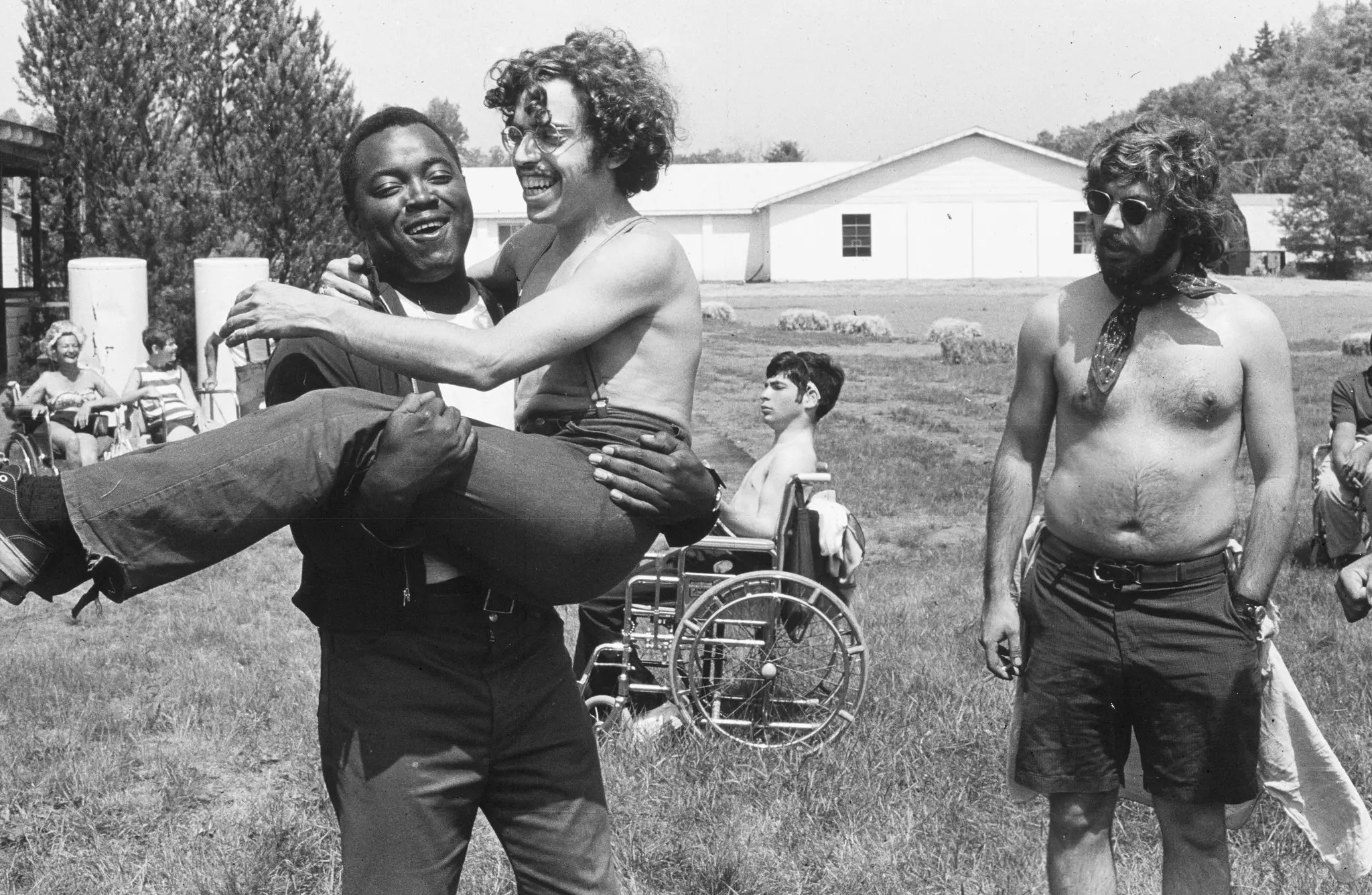 A smiling man holds another laughing man in his arms outside on a grassy field, capturing the spirit of inclusive fatherhood. In the background, people mingle, including one in a wheelchair and a shirtless man with sunglasses and a scarf.