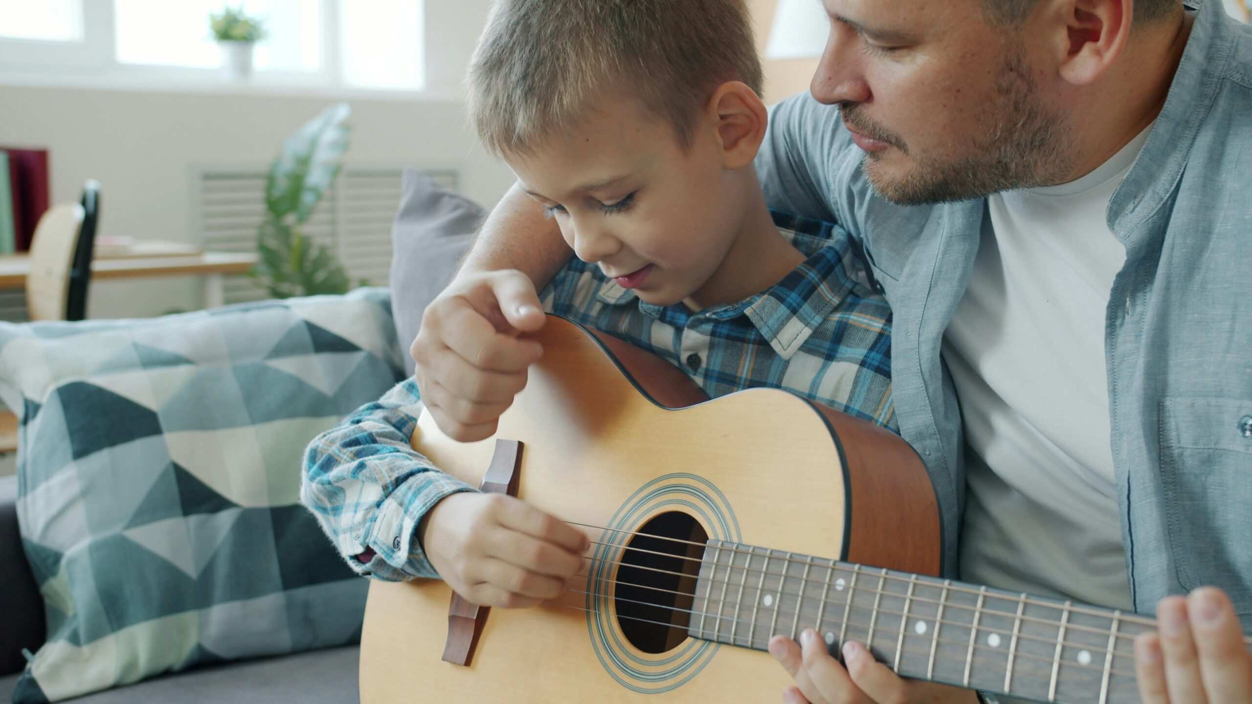 A man helps a young boy play an acoustic guitar, guiding his hand on the strings. They are sitting closely together on a couch in a bright living room with patterned pillows.