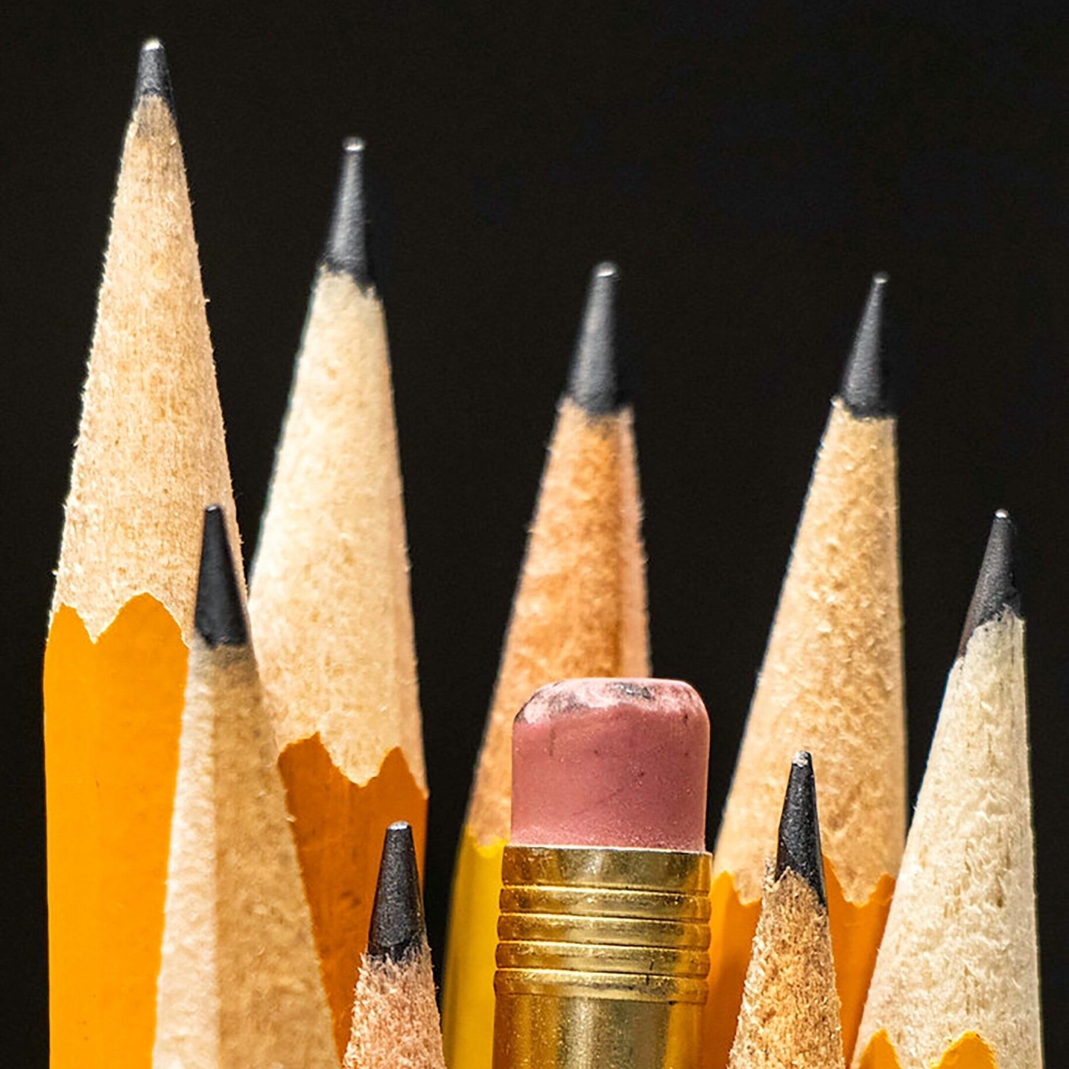 Close-up of several sharpened yellow pencils with one pencil, showing a used pink eraser, positioned upside down among them. The background is dark, highlighting the pencils points and textures.