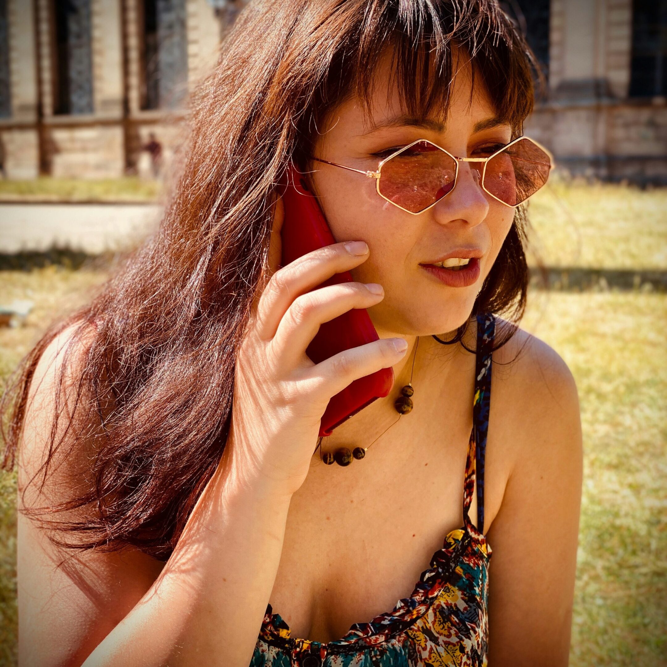 A woman with long brown hair and rose-tinted sunglasses is outdoors, holding a red smartphone to her ear. She is wearing a sleeveless floral dress and a beaded necklace, with sunlight highlighting her face.