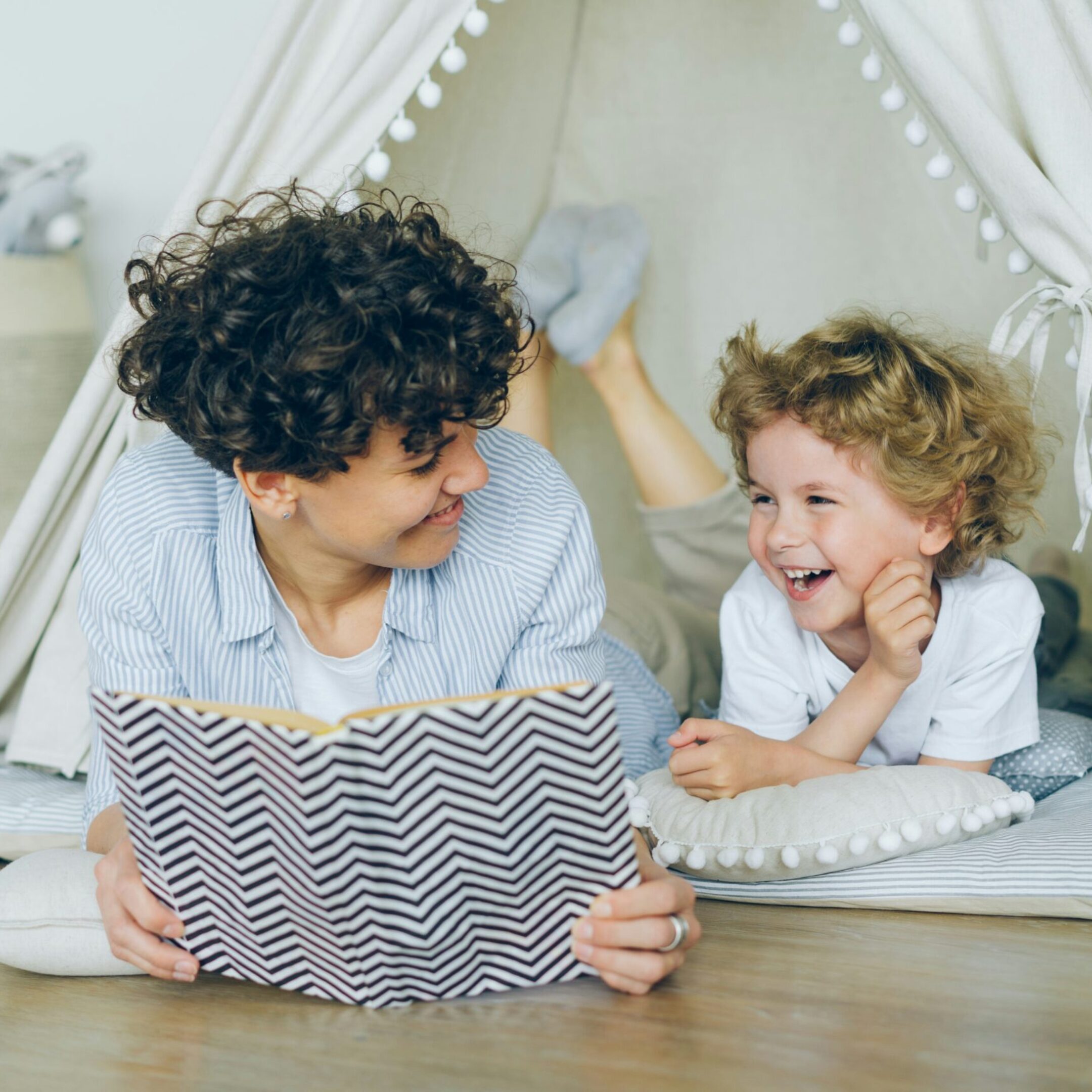 An adult and a child with curly hair smile at each other while lying on the floor inside a cozy play tent. The adult holds an open book, and the child rests on a pillow, both looking happy and engaged.
