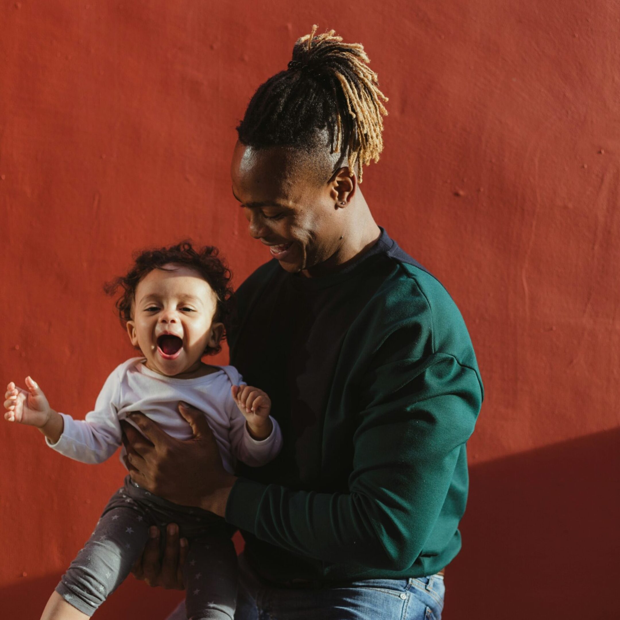 A smiling man holding a happy toddler in his arms, standing against a red wall. The child is laughing with arms outstretched, and sunlight casts soft shadows on them.